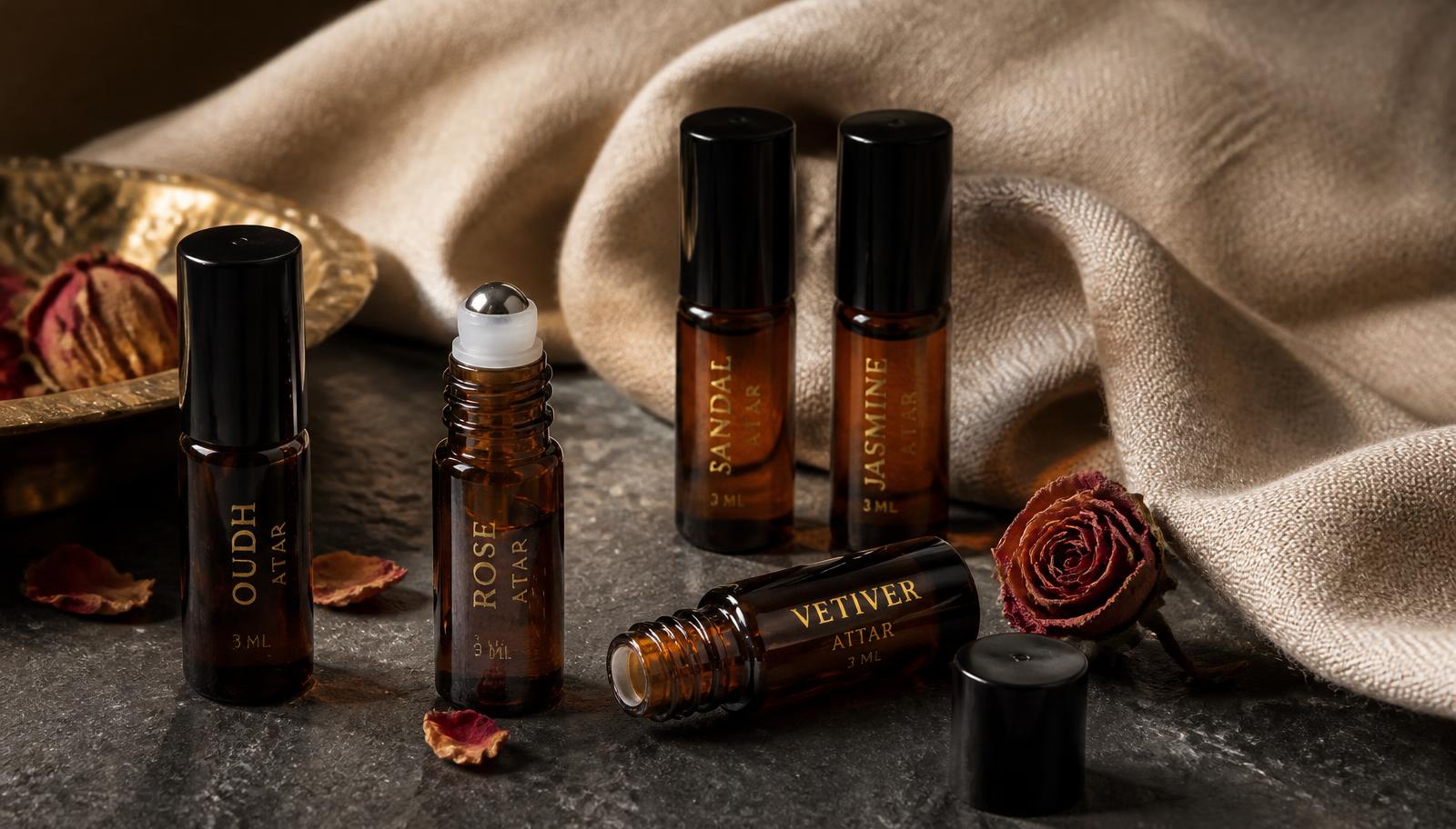 Five amber glass attar roll-on bottles on dark stone with linen cloth and dried roses